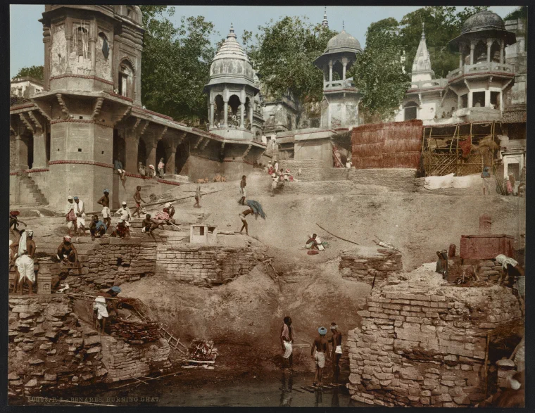 Colour photochrom of a crowded riverside ghat at Benares with stone steps, buildings, and boats on the Ganges