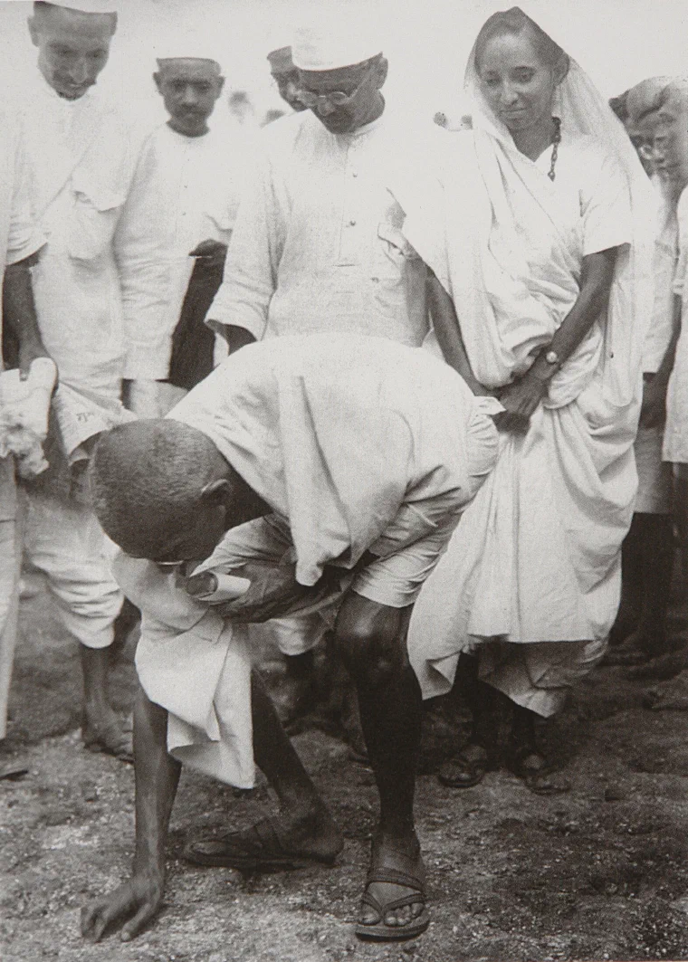 Photograph of Gandhi stooping at the shoreline at Dandi on 5 April 1930, surrounded by followers