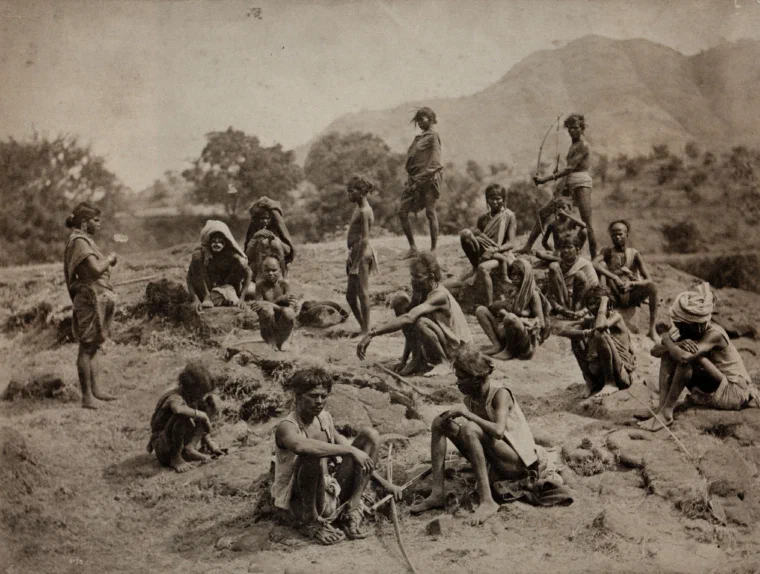 Sepia nineteenth-century photograph of a Bhil group posed outdoors on rocky ground with hills behind them