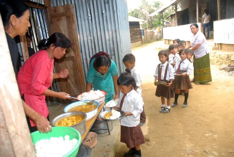 Children seated on a school veranda in Wokha district, Nagaland, as adults serve a cooked midday meal